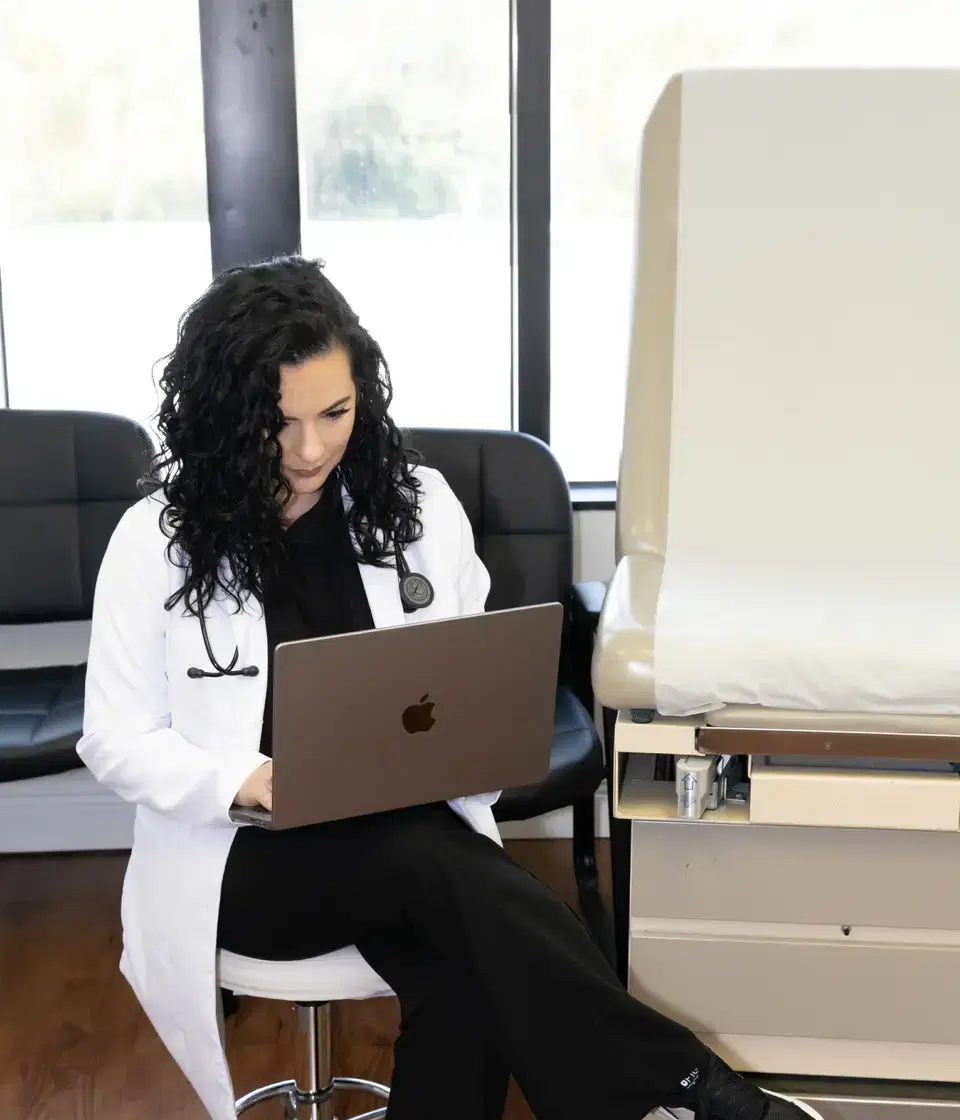 Una médica con bata blanca y estetoscopio atiende una consulta desde su computadora portátil en un consultorio.
